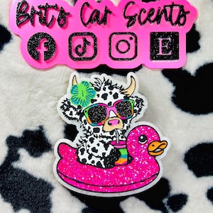May include: A pink car scent charm with the text "Brit's Car Scents" and social media icons. Below is a cow wearing sunglasses and floating on a pink duck floatie, holding a drink. The background is a black and white cow print.