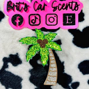May include: A pink car scent logo with the text "Brit's Car Scents" and social media icons. Below is a glittery green palm tree with a brown trunk and glittery coconuts. The background is a black and white cow print.