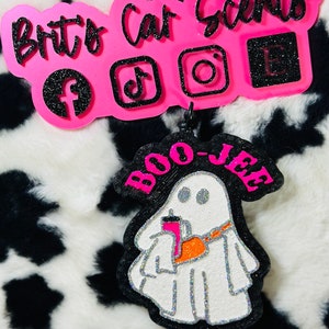 K&ouml;nnte beinhalten: Ein dekorativer Auto-Lufterfrischer mit einem wei&szlig;en, glitzernden Geist, der einen pinken Becher h&auml;lt und eine orangefarbene Umh&auml;ngetasche tr&auml;gt, mit dem Text BOO-JEE in Pink. Dar&uuml;ber zeigt ein pinkes Schild Brit's Car Scents mit Social-Media-Symbolen.