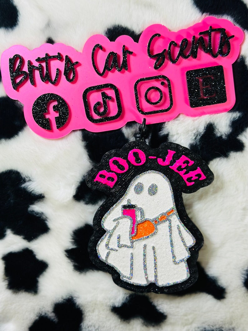 K&ouml;nnte beinhalten: Ein dekorativer Auto-Lufterfrischer mit einem wei&szlig;en, glitzernden Geist, der einen pinken Becher h&auml;lt und eine orangefarbene Umh&auml;ngetasche tr&auml;gt, mit dem Text BOO-JEE in Pink. Dar&uuml;ber zeigt ein pinkes Schild Brit's Car Scents mit Social-Media-Symbolen.