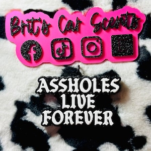 May include: Pink and black car air freshener with the text "Britt's Car Scents" and social media icons. The air freshener also has the text "Assholes Live Forever" in white letters on a black background.