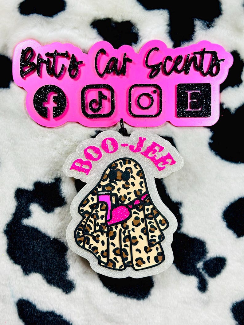 K&ouml;nnte beinhalten: Ein dekorativer Auto-Lufterfrischer in Form eines Geistes mit Leopardenmuster, der eine pinke Umh&auml;ngetasche tr&auml;gt und einen pinken Becher h&auml;lt. Dar&uuml;ber befindet sich ein pinkes Schild mit der Aufschrift Brit's Car Scents und Social-Media-Symbolen.