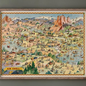 Op de afbeelding: Een ingelijste vintage kaartillustratie met een gedetailleerde, kleurrijke weergave van een landschap met mensen, voertuigen en bezienswaardigheden. De tekst "ASK SHELL!" is zichtbaar. Het frame is van hout.