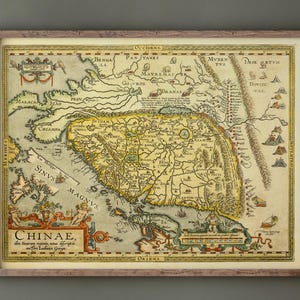 Può includere: Una mappa antica incorniciata della Cina, con dettagli intricati e un'estetica vintage. La mappa è prevalentemente beige e gialla, con testo in latino e un bordo decorativo. La cornice è di colore marrone chiaro.