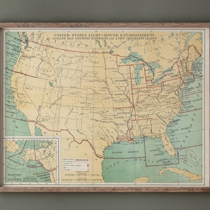 May include: A framed vintage map of the United States, showing light-house districts. The map features a detailed outline of the country with red lines and text, set against a beige background. The frame is made of light-colored wood.