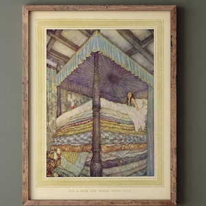 Op de afbeelding: Een ingelijste prent van een sprookjesillustratie. Het kunstwerk toont een vrouw in een bed volgepakt met kleurrijke matrassen onder een baldakijn. De tekst onderaan luidt "NOT A WINK THE WHOLE NIGHT LONG."