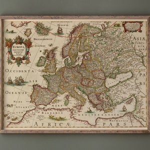 Op de afbeelding: Een ingelijste antieke kaart van Europa op crèmekleurig perkamentpapier met gedetailleerde cartografie, decoratieve illustraties van schepen en sierlijke tekstlabels waaronder EVROPA EXACTISSIME DESCRIPTA in een bruine houten lijst.
