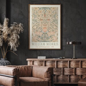 K&ouml;nnte beinhalten: Ein gerahmter Druck einer Vintage-botanischen Illustration von William Morris, mit einem sich wiederholenden Muster aus Lilien und Granat&auml;pfeln in Gr&uuml;n-, Rosa- und Beiget&ouml;nen. Der Titel "Lily and Pomegranate - 1886" ist unter dem Bild gedruckt.