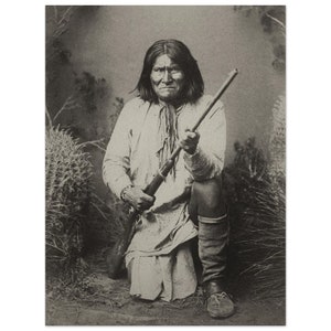 Könnte beinhalten: Ein Schwarzweißfoto eines indianischen Mannes, der ein Gewehr hält. Er trägt ein langärmeliges Hemd und eine Hose. Er kniet in einem Feld mit hohem Gras.