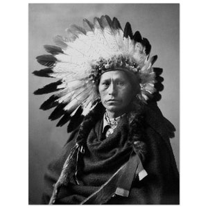 May include: A black and white portrait of a Native American man wearing a traditional headdress with feathers. He is wearing a dark colored blanket and has a serious expression on his face.