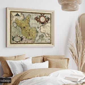 May include: A framed vintage map with a light wood frame. The map features a detailed illustration with shades of yellow, brown, and blue. Ornate cartouches and text are visible. The map is displayed above a bed with neutral-toned bedding.