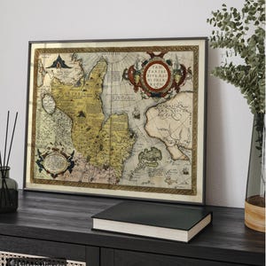 May include: Framed antique map with a detailed depiction of a historical region. The map features intricate cartography with various text elements and decorative borders. The frame is dark, and the map is displayed on a dark wooden surface with a book and a vase.