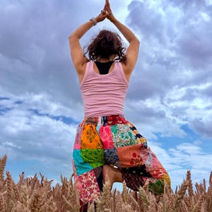 May include: A pair of loose-fitting patchwork pants featuring a vibrant mix of multi-colored floral and geometric fabric squares, paired with a light pink tank top, worn while standing in a field of wheat under a cloudy blue sky.
