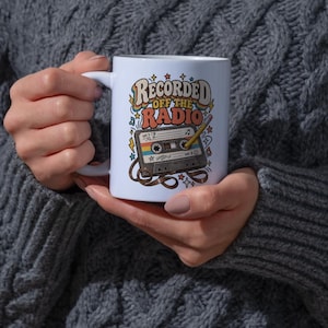 May include: A white ceramic mug featuring a colourful graphic of a cassette tape with a pencil inserted into the spool. The text reads "RECORDED OFF THE RADIO" in a retro font, surrounded by small stars, musical notes, and lightning bolt icons.