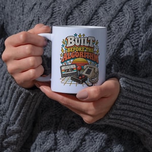May include: A white ceramic mug featuring a colorful graphic with the text "BUILT BEFORE THE ALGORITHM" in a retro font. The design includes a cassette tape, a joystick, and a rotary telephone set against a sunset and a grid pattern background.