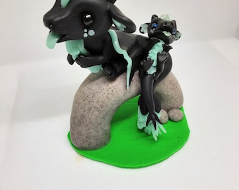Op de afbeelding: Zwarte en groene drakenfiguur met een kleinere drakenfiguur op zijn rug. Beide draken hebben lichtgroene accenten en zitten op een grijze rots met groen gras.