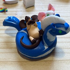 Puede incluir: Una pequeña figura de arcilla que muestra un ratón color canela con orejas y cola marrón oscuro descansando sobre un flotador circular azul y blanco. Una criatura blanca y rosa con ojos verdes está situada en la parte delantera del flotador.