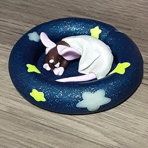 May include: A handmade figurine of a sleeping rat in a dark blue, glittery ring. The rat has brown fur, pink ears, and wears a white garment. The ring is decorated with white clouds and yellow stars.