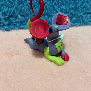 May include: A whimsical, handmade figurine of a mouse-like creature. It features a lavender body, red ears and tail, green feet and accents, and patches with designs. The figurine is set against a blue and pink background.
