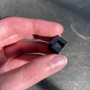 May include: A small, black, 3D printed cube with a square hole in the center.
