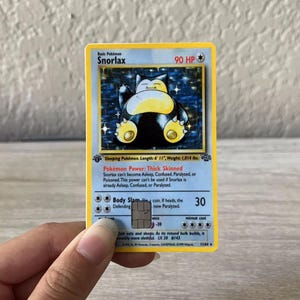 Puede incluir: Una carta coleccionable de Pok&eacute;mon Snorlax con borde azul y amarillo. La carta presenta una ilustraci&oacute;n de Snorlax durmiendo y texto que incluye "90 HP", "Body Slam" y "LV. 20 #143". La carta es sostenida por una mano.