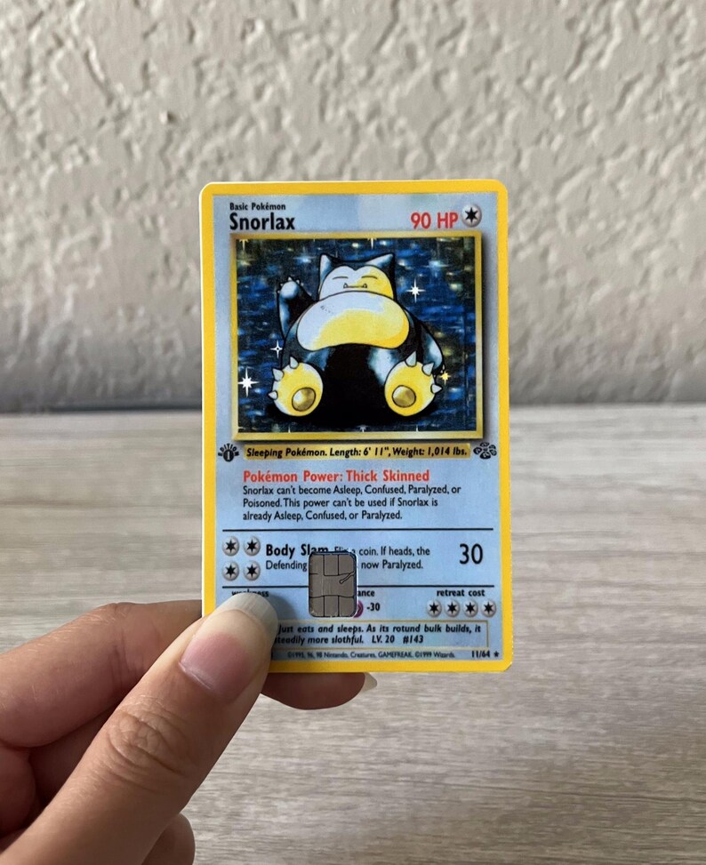 Puede incluir: Una carta coleccionable de Pok&eacute;mon Snorlax con borde azul y amarillo. La carta presenta una ilustraci&oacute;n de Snorlax durmiendo y texto que incluye "90 HP", "Body Slam" y "LV. 20 #143". La carta es sostenida por una mano.
