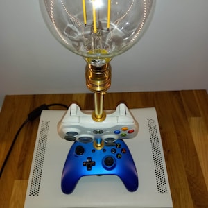 May include: A novelty lamp with a large, clear globe light bulb and a gold base. The lamp is mounted on two game controllers, one white and one blue, which are placed on top of a white game console. The lamp is plugged in.