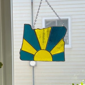 May include: A stained glass window hanging in a window, shaped like the state of Oregon, with a yellow sun rising over a teal blue background.