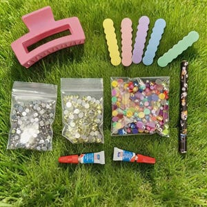 May include: A selection of craft supplies, including a pink hair claw clip, colourful barrettes, bags of rhinestones, glue tubes, and a patterned pen. The items are arranged on green grass.
