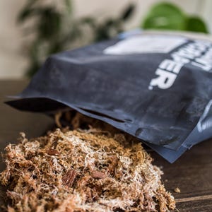 Peut inclure: Un sac noir partiellement ouvert, laissant appara&icirc;tre un tas de sphaigne brun et beige. La mousse est grossi&egrave;re et fibreuse, avec des morceaux d'&eacute;corce visibles. L'arri&egrave;re-plan est flou, avec une surface en bois.