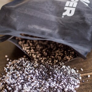 Puede incluir: Una bolsa negra de mezcla de tierra est&aacute; abierta, derramando una mezcla de tierra oscura y gr&aacute;nulos de perlita blancos sobre una superficie de madera. La bolsa tiene texto blanco. La imagen es un primer plano.