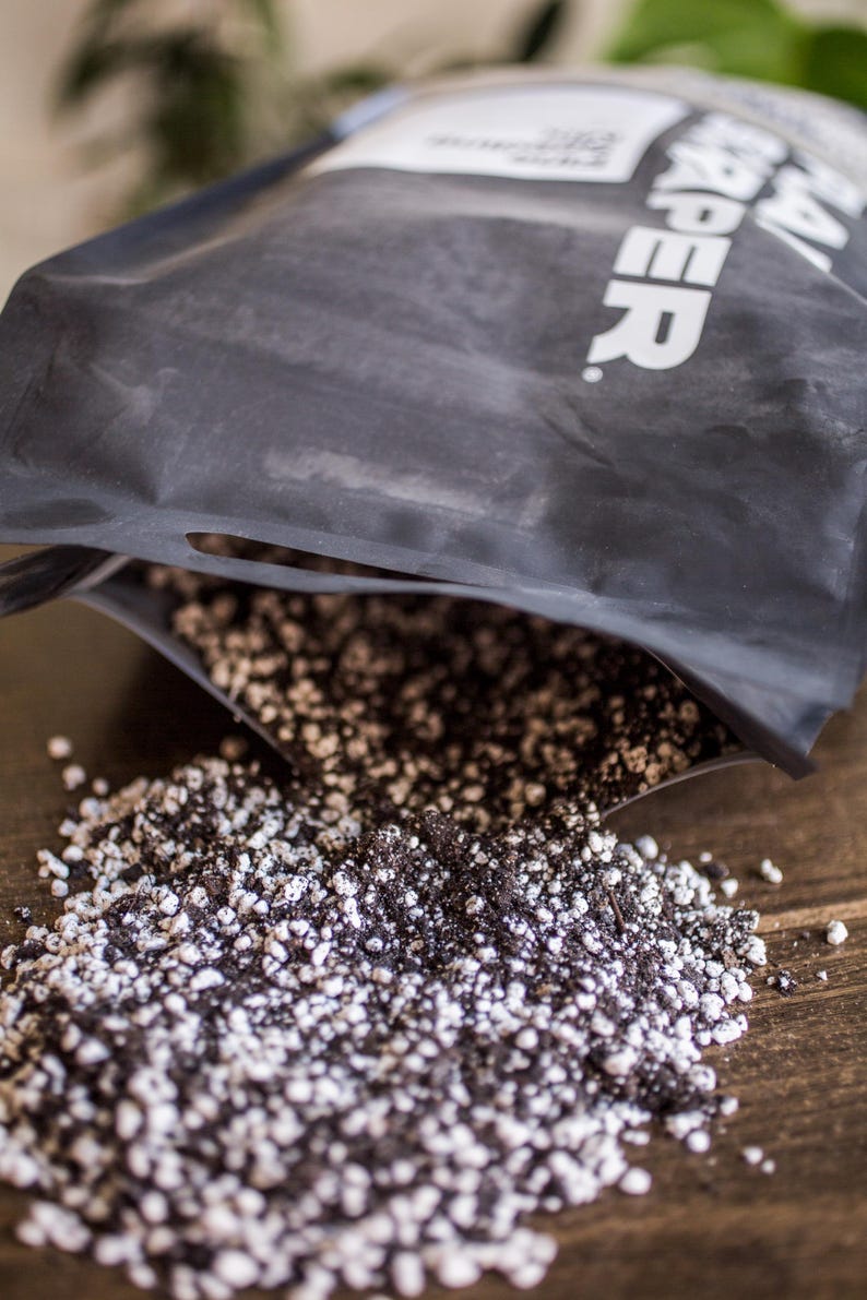 Puede incluir: Una bolsa negra de mezcla de tierra est&aacute; abierta, derramando una mezcla de tierra oscura y gr&aacute;nulos de perlita blancos sobre una superficie de madera. La bolsa tiene texto blanco. La imagen es un primer plano.