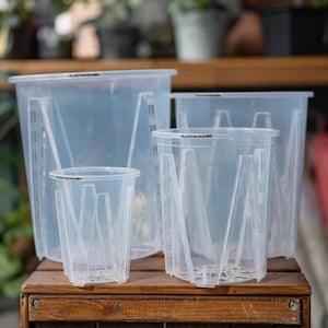 Op de afbeelding: Een verzameling doorzichtige plastic plantenpotten van verschillende formaten, gerangschikt op een houten oppervlak. De potten hebben een sleufontwerp en de tekst "PLANTSCRAPER" is op de rand gedrukt. De achtergrond toont vage groenvoorziening.