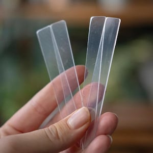Puede incluir: Una mano sosteniendo varias etiquetas de plástico transparentes y rectangulares para plantas. Las etiquetas son delgadas y transparentes, con un borde superior ligeramente redondeado. El fondo está borroso.