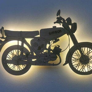 Könnte beinhalten: Holzschnitt-Silhouette eines Motorrads mit dem Wort "Simson" auf dem Tank. Das Motorrad wird von hinten beleuchtet, wodurch ein warmer Schein entsteht. Das Design umfasst Details wie Räder, Sitz und Lenker.