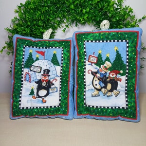 May include: Two blue and green fabric pot holders with a winter theme. One features a penguin with a sign that says "Greatest Show on Ice" and the other features two penguins ice skating with a sign that says "Fun on Ice Today".