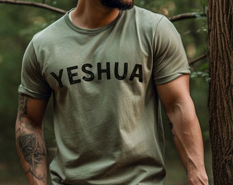 May include: A man wearing a green t-shirt with the word "YESHUA" printed in black letters. He is standing in a forest setting.