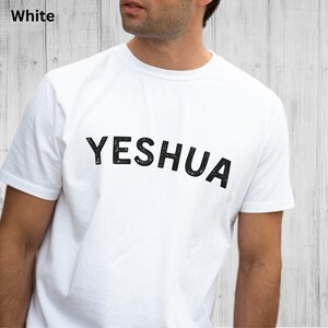 以下が含まれることがあります： 前面に黒い文字で「YESHUA」とプリントされた白いTシャツです。