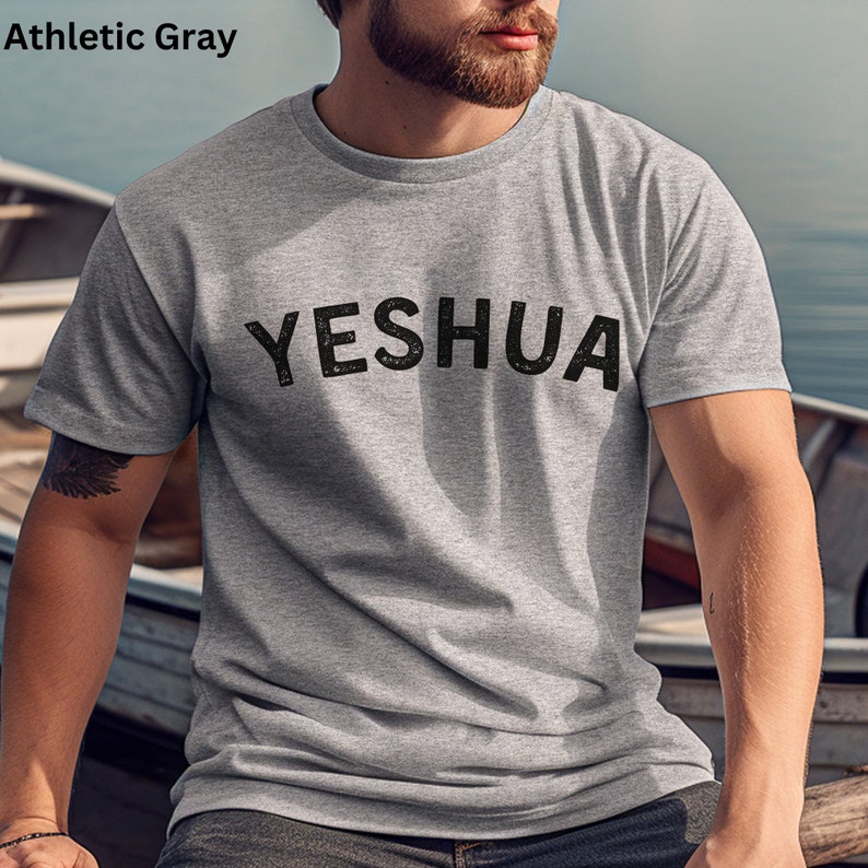 以下が含まれることがあります： 黒い文字で「YESHUA」とプリントされたグレーのTシャツです。