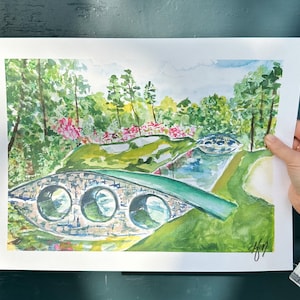May include: Watercolor painting of a golf course with a green bridge over a stream. The course is surrounded by trees and bushes, and there is a small pond in the background. The painting is signed in the bottom right corner.