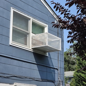 Puede incluir: Un recinto rectangular blanco para gatos, hecho de un marco de metal y malla, que se extiende hacia afuera desde la ventana de una casa con revestimiento azul. La estructura tiene forma de caja con una parte superior plana y lados de malla.