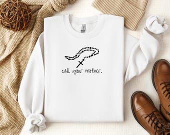 May include: White crew neck sweatshirt with a black rosary and cross design. The phrase "call your mother" is printed below the rosary. The sweatshirt is displayed with a beige beanie and brown leather boots.