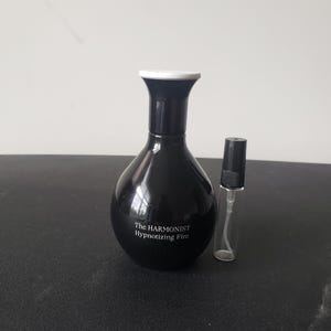 May include: A black glass bottle of perfume with a white cap. The bottle is labeled "The Harmonist Hypnotizing Fire". A smaller, clear glass bottle with a black cap is also pictured.