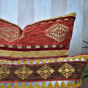 Puede incluir: Dos almohadas decorativas con un patrón geométrico en tonos rojos, marrones, dorados y plateados. Las almohadas están hechas de una tela suave y texturizada.