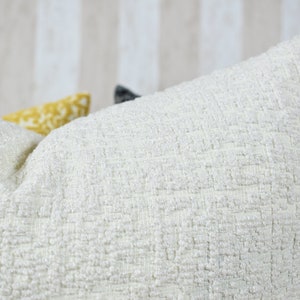 May include: A set of three decorative throw cushions. The cushions are made from a textured fabric and feature a variety of colours and patterns. The cushions are arranged in a row, with the white cushion at the front.