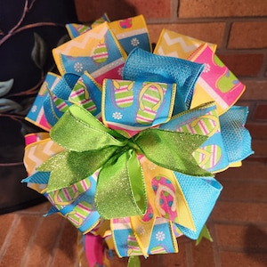 Puede incluir: Un lazo decorativo con cintas coloridas con diseños de chanclas en azul, amarillo y rosa. El lazo está acentuado con un gran lazo verde brillante en el centro, perfecto para decoraciones de verano o temáticas de playa.