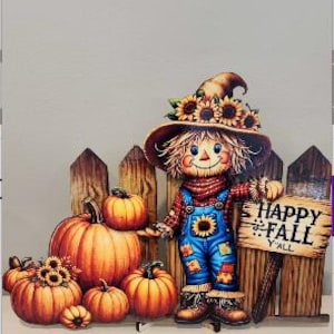 May include: A decorative cutout featuring a scarecrow wearing a hat with sunflowers, blue overalls, and a red scarf, standing next to a wooden fence with pumpkins and a sign that reads HAPPY FALL Y'ALL. The scene includes several orange pumpkins.