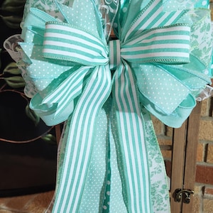 May include: A large decorative bow featuring various ribbons in shades of turquoise and white. The bow includes ribbons with polka dots, stripes, and floral patterns. The bow is attached to a wooden frame.