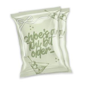Puede incluir: Dos bolsas de patatas fritas verdes y blancas con una cinta blanca y el texto "Chloe's a tiny bit older..." impreso en la parte delantera. Las bolsas están decoradas con una copa de martini con aceitunas y estrellas.