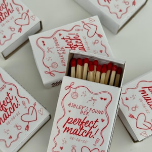 May include: White matchboxes with red hearts and illustrations. The text on the boxes reads "Ashley's found her perfect match!" with the date "06-06-25".